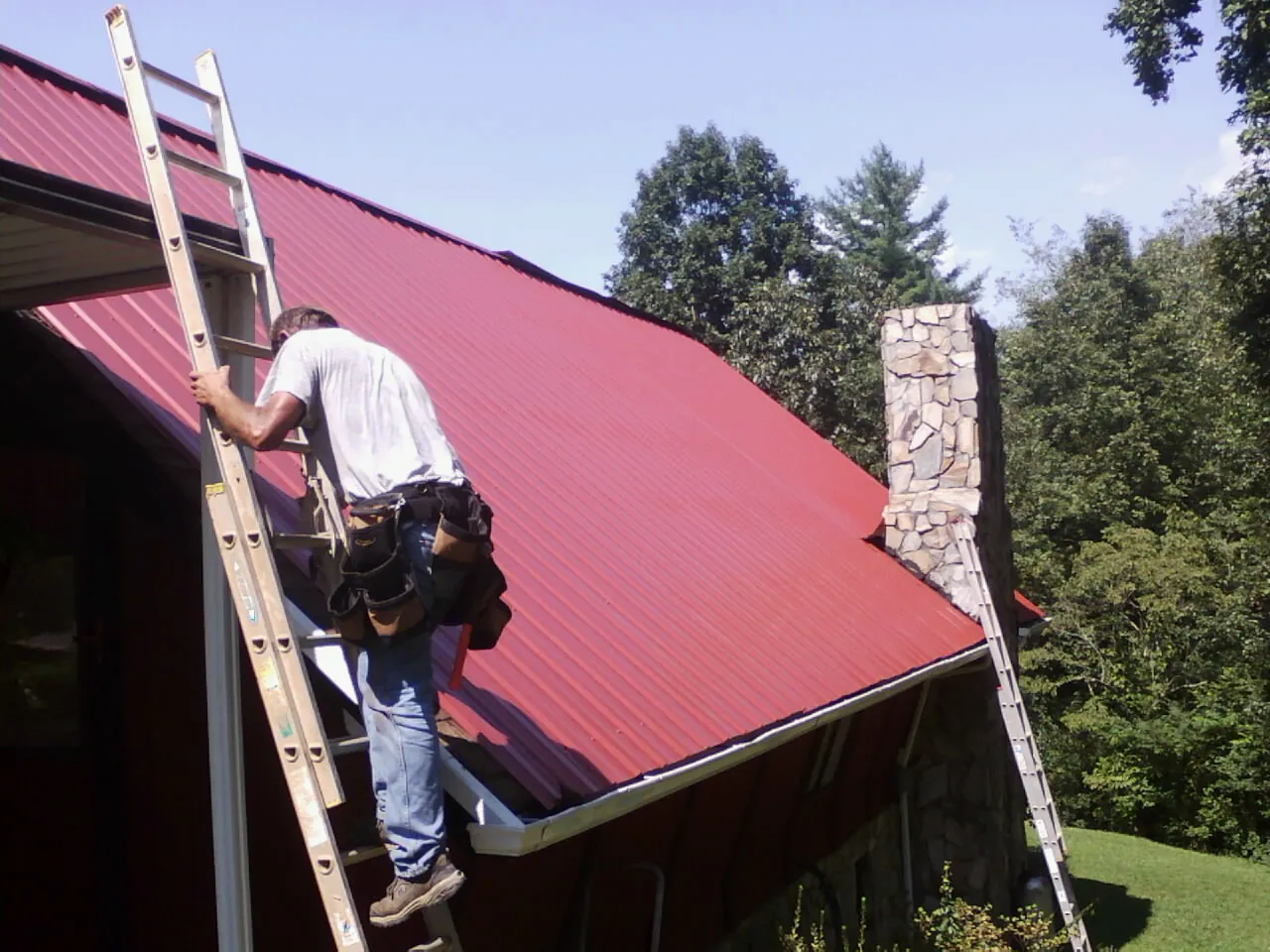 Roofer on a ladder beside a red metal roof and stone chimney — Soffit & Fascia Repair by Roofing Brothers in Turon